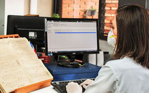 Foto de funcionaria frente a computadora realizando descripción de documento antiguo