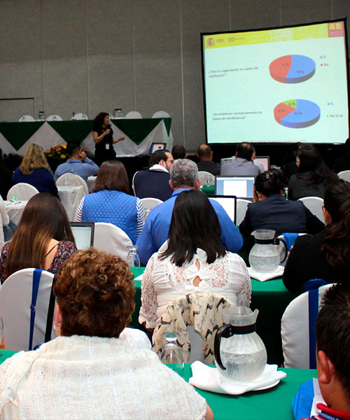 Foto de personas recibiendo charla en salón de eventos, al fondo la expositora